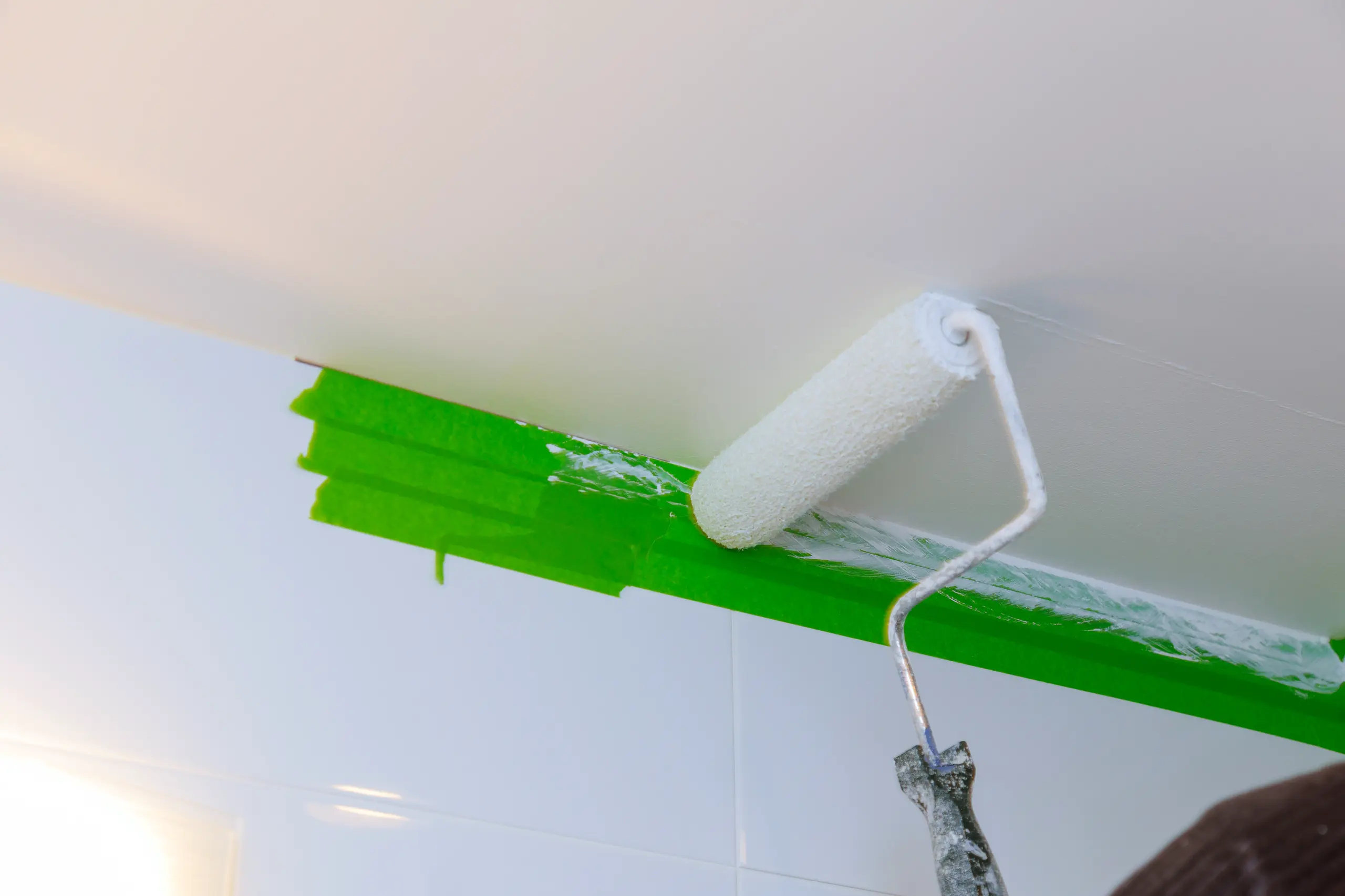 Close-up of a paint roller applying ceiling paint with green masking tape in the background